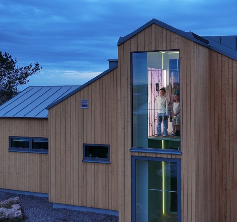 Bild zu Aufzughersteller "Swift" aus Schweden Blick von außen in ein modernes Holzhaus mit einem gläsernen Fahrstuhl, darin ein Erwachsener und zwei Kinder